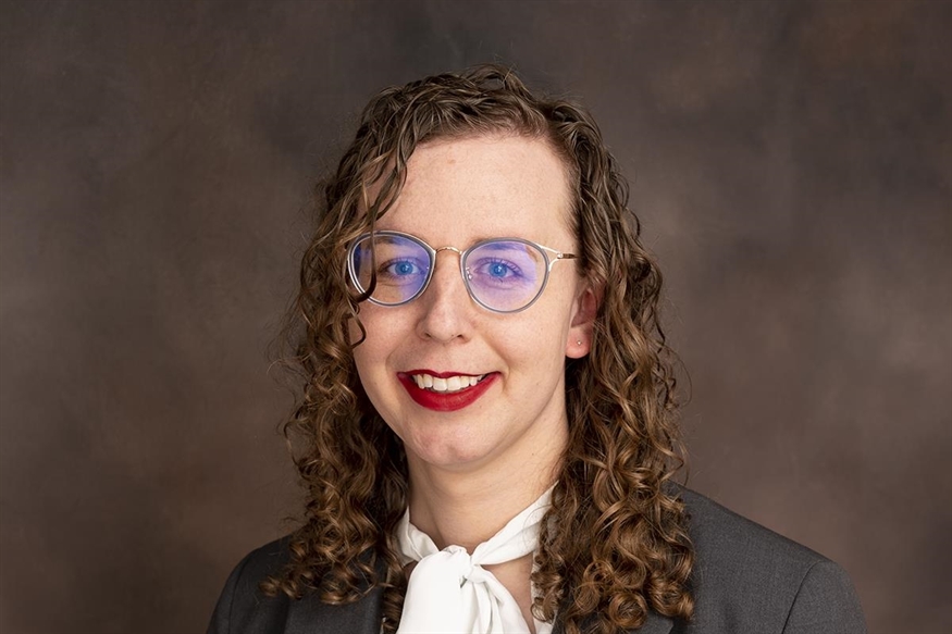 [ai] A person with curly hair and glasses smiling while dressed in a suit and a white scarf. The background is a muted brown tone.